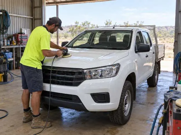 An operator machine polishing the bonnet of a white dual cab ute inside an open workshop shed