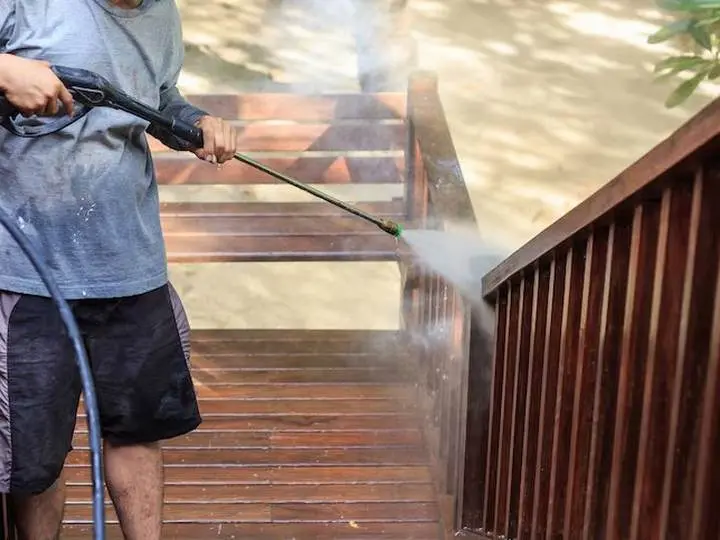 An operator washing a timber deck with a pressure wand, spray visible in the sunlight